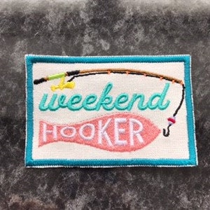 Pode incluir: Um remendo de tecido branco com uma borda turquesa. O remendo apresenta um peixe rosa com o texto "weekend hooker" em letras turquesa. Uma vara de pesca amarela e marrom com um anzol está acima do peixe.