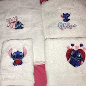 Puede incluir: Conjunto de cuatro toallas de baño blancas con personajes bordados de Stitch y Angel de la película de Disney Lilo & Stitch. Las toallas incluyen una toalla de mano con Stitch sosteniendo un corazón, una toalla de baño con Stitch y Angel, una toalla de baño con Stitch y la palabra "Ohana" y una toalla de baño con Stitch en un corazón con huellas de patas rosas.