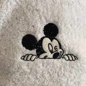 May include: Black and white embroidered image of Mickey Mouse peeking over a white surface. Mickey Mouse is looking to the right with a mischievous expression.