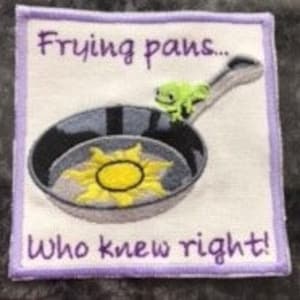 Op de afbeelding: Een vierkante patch in paars en wit met een groene kikker die op een koekenpan zit. De koekenpan heeft een gele zonnebloem erin. De tekst op de patch luidt "Frying pans... who knew right!"