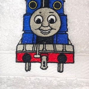 May include: Embroidered blue and red cartoon train engine with a smiling face, black eyes, and yellow wheels. The train is on a white background.