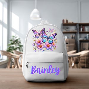 Peut inclure: Sac à dos blanc orné d'un motif de papillons et de fleurs colorées. Il comporte une poche avant et le nom "Brinley" en dégradé de violet. Les papillons ont des ailes bleues, violettes et roses.
