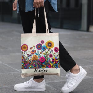 Puede incluir: Un bolso tote de lona con un diseño floral colorido. Las flores son en tonos de rosa, amarillo, azul y morado. El nombre "Brinley" está impreso en escritura negra en la parte inferior de la bolsa.