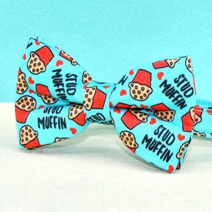 May include: A light blue cat collar and bow tie set. The bow tie and collar feature a repeating pattern of red muffin cups and the words "Stud Muffin". The set includes a gold bell and a white pearl bell.
