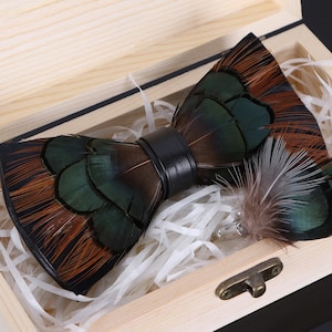 Puede incluir: Una pajarita de plumas marrón, verde y negra, presentada en una caja de madera clara con relleno de papel. La pajarita tiene una banda de cuero negro. También se incluye un pin de solapa de plumas a juego.