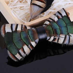 Puede incluir: Una pajarita de plumas hecha a mano con una banda de cuero negro, con plumas verdes, marrones y blancas. Un pin de solapa de plumas a juego está en una caja de madera con relleno de papel. Un accesorio único.