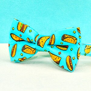 May include: A turquoise cat bow tie and collar set with a repeating pattern of yellow and orange tacos. The collar has a gold bell and a white underside. The bow tie is centered in the image.