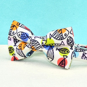 May include: A white cat collar and bow tie set with a colorful fish pattern. The bow tie features a variety of fish illustrations in black, orange, blue, and green, set against a white background. The collar has a gold bell.
