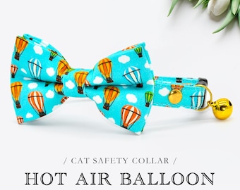 May include: A turquoise cat safety collar and bow tie set, patterned with hot air balloons in orange, yellow, white, and green. The collar has a gold bell and buckle. The image text reads "HOT AIR BALLOON".