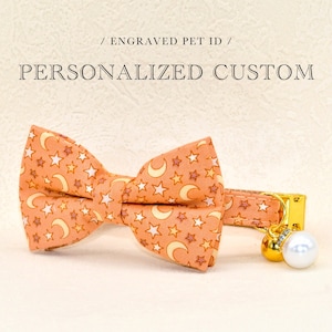 May include: A peach-colored cat collar with a matching bow tie featuring a crescent moon and star pattern. The collar has a gold bell with a pearl charm.