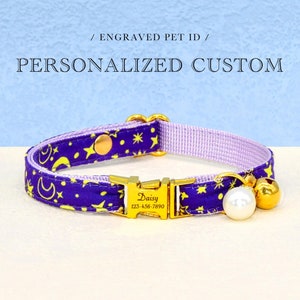 Peut inclure: Un collier personnalisé pour animaux de compagnie avec un motif céleste violet et jaune. La boucle dorée affiche le nom et le numéro de téléphone de l'animal. Une perle blanche et une cloche dorée sont attachées. Texte: / Engraved Pet ID / PERSONNALISÉ SUR MESURE.