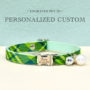 May include: A personalized pet collar with a green and blue plaid pattern. The collar has a silver buckle engraved with a name and phone number. A silver bell and a white pearl charm dangle from the collar. The text "PERSONALIZED CUSTOM" is above the collar.