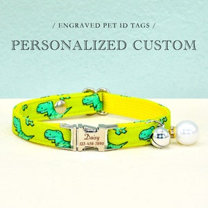 May include: Yellow cat collar with a green dinosaur print and a silver engraved tag that reads "Daisy 123-456-7890". The collar has a silver bell and a white pearl charm.
