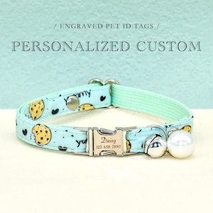 Könnte beinhalten: Ein hellblaues Katzenhalsband mit einem Keksmuster und einer silbernen Schnalle. Die Schnalle hat ein personalisiertes, graviertes Schild mit der Aufschrift "Daisy 123-456-7890". Das Halsband hat außerdem eine silberne Glocke und einen weißen Perlenanhänger.