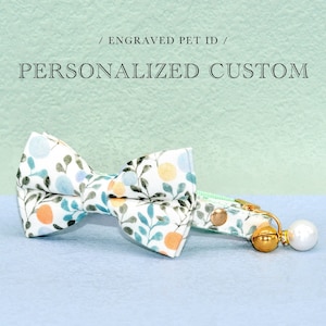 Könnte beinhalten: Ein weißes Katzenhalsband mit einer grünen und orangefarbenen Schleife mit Blumenmuster und einer goldenen Glocke mit einem Perlenanhänger. Das Halsband trägt den Text "ENGRAVED PET ID / PERSONALIZED CUSTOM" auf grünem Hintergrund.