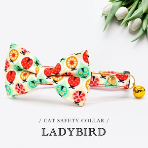 May include: A red, white and green cat collar with a ladybug print and a gold bell. The collar has a bow tie attached. / CAT SAFETY COLLAR / LADYBIRD