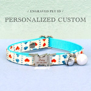 May include: A personalized custom pet collar with a white and patterned fabric featuring red hearts and blue spades. The collar has a turquoise blue backing and a silver metal buckle engraved with a name and phone number. A silver bell and white pearl charm are attached.