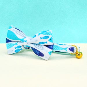 May include: A pet collar and bow tie set with a fish pattern. The bow tie is white with blue fish illustrations. The collar has a gold-coloured bell and a matching fish design.