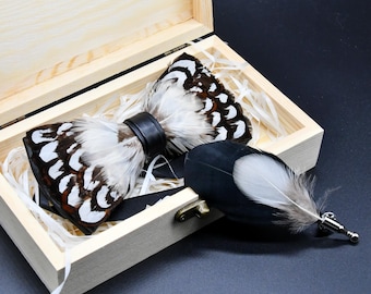 May include: A feather bow tie with a black leather band, presented in a wooden box with a matching feather lapel pin. The bow tie features white, brown, and black feathers. The box is lined with white paper.