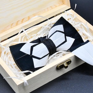 May include: A black and white patterned bow tie with a geometric design, presented in a wooden box with decorative paper. The bow tie is secured with a black cord. The box has a metal clasp.