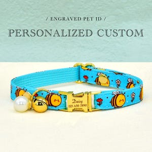 May include: A light blue pet collar with a bee and flower pattern. The collar has a gold-colored buckle engraved with a name and phone number. A white pearl and a gold bell are attached to the collar. The text "Engraved Pet ID" and "Personalized Custom" are in the background.