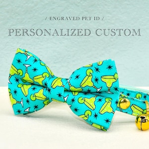 May include: A turquoise pet bow tie with a margarita pattern and a gold bell. The bow tie has the text "/ ENGRAVED PET ID / PERSONALIZED CUSTOM". The bow tie is designed for a cat or dog.