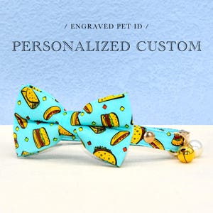May include: A blue cat collar with a bow tie featuring a pattern of yellow tacos and hamburgers. The collar has a bell and a gold buckle.