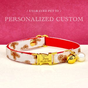 May include: A personalized pet collar with a white fabric featuring gingerbread men and red backing. The gold buckle is engraved with "Daisy" and a phone number. A gold bell and pearl charm hang from the collar. The text reads "/ ENGRAVED PET ID / PERSONALIZED CUSTOM".