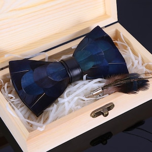 May include: A navy blue feather bow tie with a black leather band, presented in a light wooden box with white paper filler. A matching feather lapel pin is included. The bow tie has a unique, textured appearance.