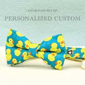May include: A blue pet bow tie with a yellow duck pattern. The bow tie is part of a collar with a metal clasp. The text "/ ENGRAVED PET ID / PERSONALIZED CUSTOM" is above the bow tie.