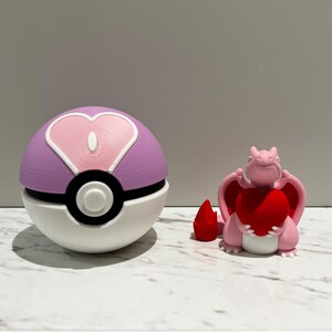 May include: A pink and white Poke Ball with a pink heart design on the top half and a black and white Poke Ball design on the bottom half. A pink dragon figurine holding a red heart is next to the Poke Ball.