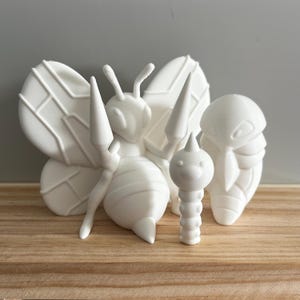 May include: A set of white 3D-printed insect models, featuring a large bee with detailed wings, a small caterpillar, and a cocoon shape. The figures are displayed on a wooden surface, set against a grey background.