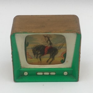 May include: A small, rectangular decorative object shaped like a vintage television set. It features a brown wooden-look frame, a bright green front panel with four small white buttons, and a screen displaying a graphic of a person riding a horse.