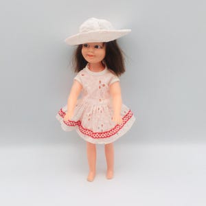 May include: A doll with brown hair wearing a white eyelet dress featuring a red scalloped trim along the hem and a matching white sun hat. The doll has painted facial features, including brown eyes and pink lips, and stands in an upright position.