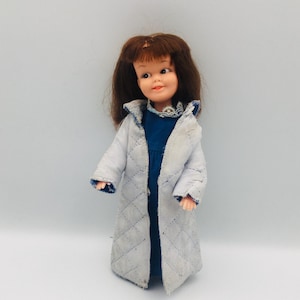 May include: A vintage doll with brown hair and a light blue quilted coat over a blue dress. The doll has a smiling expression and is standing against a white background. The coat has a diamond pattern.