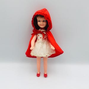 May include: A small doll with dark hair wearing a white dress with red polka dots and a scalloped hem. The doll is dressed in a bright red hooded cape that ties at the neck with a red ribbon and is wearing matching red shoes on its feet.