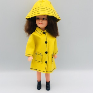 Peut inclure: Une poupée aux cheveux bruns portant un imperméable jaune vif avec des coutures noires, des boutons noirs et un chapeau de pluie jaune assorti avec des coutures noires. La poupée porte également des bottes noires, complétant la tenue.