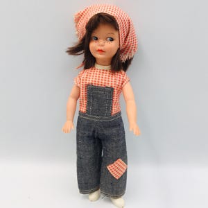 May include: A vintage doll wearing a red and white checkered headscarf and matching short-sleeved shirt. The doll has dark hair and freckles, and is wearing denim overalls with a red and white checkered patch on the knee. White shoes complete the outfit.