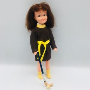 May include: A vintage doll in a brown dress with a yellow belt and collar, yellow boots, and brown hair. The doll holds a blue and white hockey stick with a ball. The doll is approximately 28 cm tall.