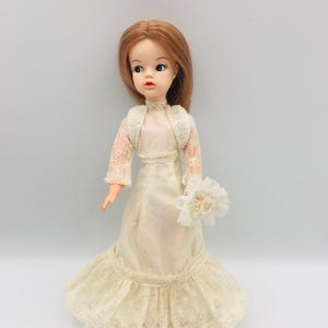 Puede incluir: Una muñeca con cabello castaño rojizo hasta los hombros, ojos azules y labios rojos. La muñeca viste un vestido color crema con corpiño de encaje, mangas largas de encaje y un dobladillo con volantes. Sostiene una flor de tela blanca.