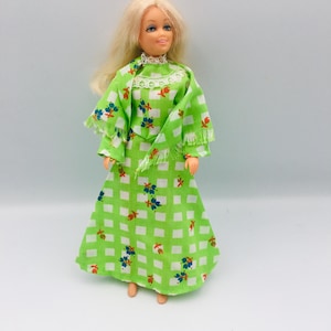 Puede incluir: Una muñeca vintage con pelo largo y rubio que lleva un vestido verde con un estampado de cuadros blancos y detalles florales. El vestido tiene mangas largas y un chal a juego con flecos. El vestido de la muñeca mide unos 30 cm de largo.