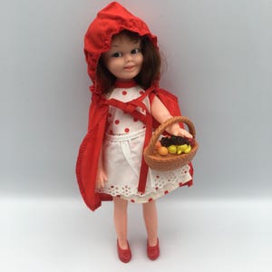 May include: A vintage doll dressed as Little Red Riding Hood, wearing a red hooded cape and a white dress with red polka dots. The doll carries a small basket filled with miniature fruits. Red shoes complete the outfit.