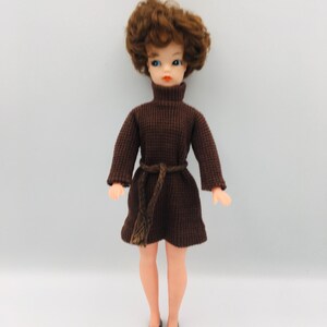 May include: A doll with short, wavy brown hair, blue eyes, and orange lips. The doll wears a dark brown, long-sleeved, ribbed turtleneck dress with a matching braided tie belt at the waist and simple black shoes. The doll has light skin.