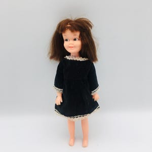 May include: A doll with shoulder-length brown hair and a painted face featuring brown eyes and pink lips. The doll wears a black long-sleeved dress with a white lace trim along the neckline, the cuffs of the sleeves, and the bottom hem of the skirt.