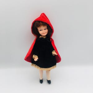 May include: A doll with brown hair wearing a black dress with gold lace trim at the sleeves and hem, paired with a bright red hooded cape that ties at the neck. The doll has light skin, painted facial features, beige stockings, and black shoes.