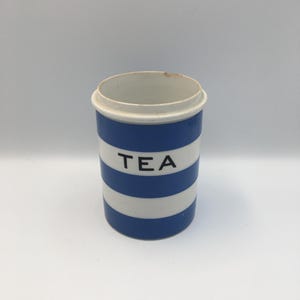 Può includere: Un contenitore per tè cilindrico in ceramica con strisce orizzontali blu e bianche. La parola TEA è stampata in lettere maiuscole nere sulla striscia bianca centrale. Il contenitore ha un interno bianco e un piccolo segno marrone sul bordo.
