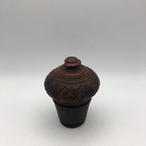 Peut inclure: Une petite boîte en bois sculptée de manière complexe avec un couvercle. La boîte est de couleur brun foncé et présente un motif géométrique sculpté sur la surface. Le couvercle est légèrement bombé et possède un petit bouton sur le dessus.