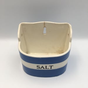May include: A wall-mounted ceramic salt cellar featuring a semi-circular shape with a flat back. The exterior is decorated with horizontal blue and white stripes, and the word SALT is printed in black capital letters on the front center.