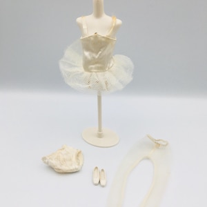 May include: A miniature ballet costume set, including a cream-colored tutu, leotard, tights, and ballet shoes. The tutu is displayed on a small mannequin. The set is designed in a neutral color palette, with a focus on delicate textures and details.