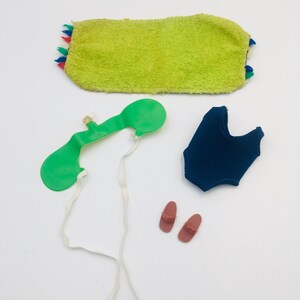 May include: A set of doll accessories including a lime green rectangular towel with multicolored fringe, a green plastic visor with white elastic straps, a dark blue one-piece swimsuit, and a pair of small brown plastic sandals.